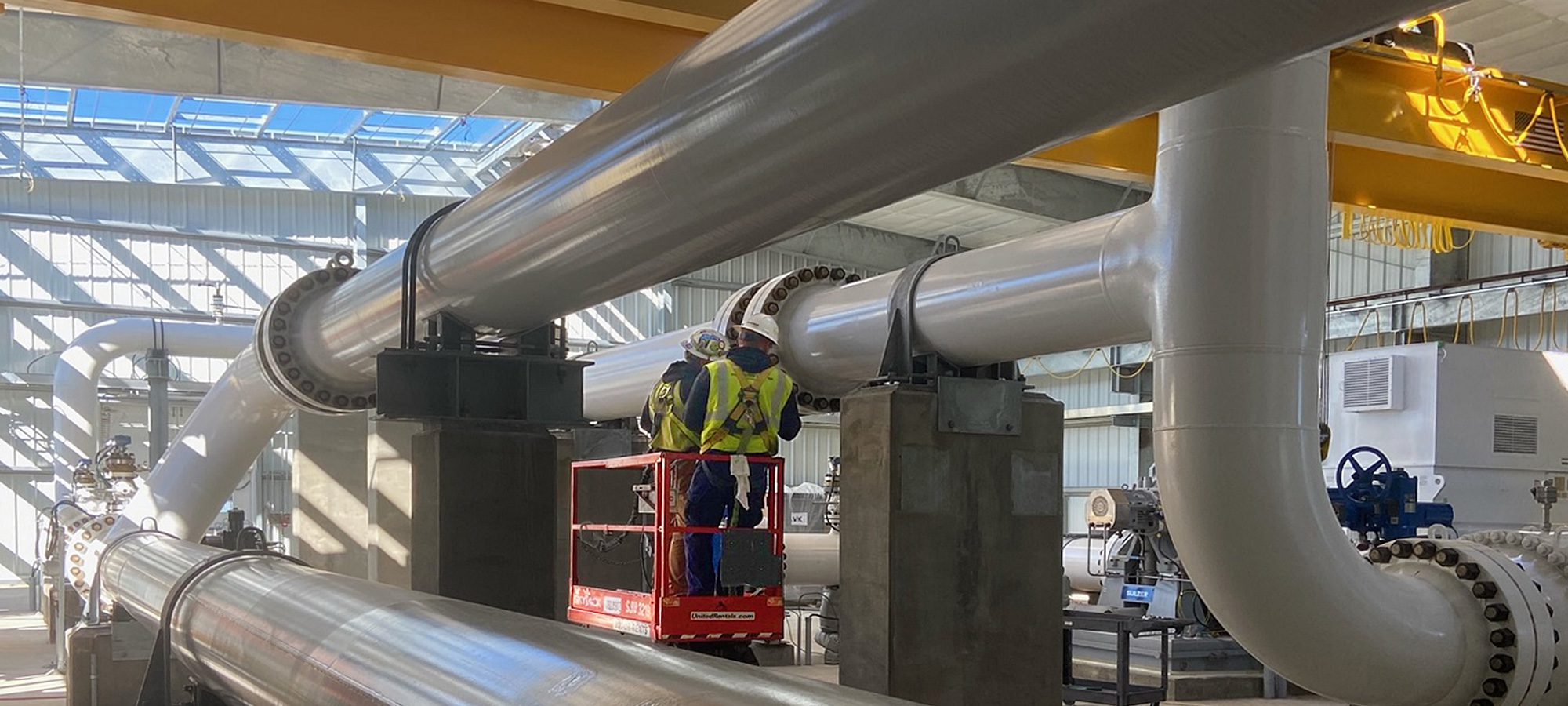large gray pipes and valves, men in safety gear on a lift